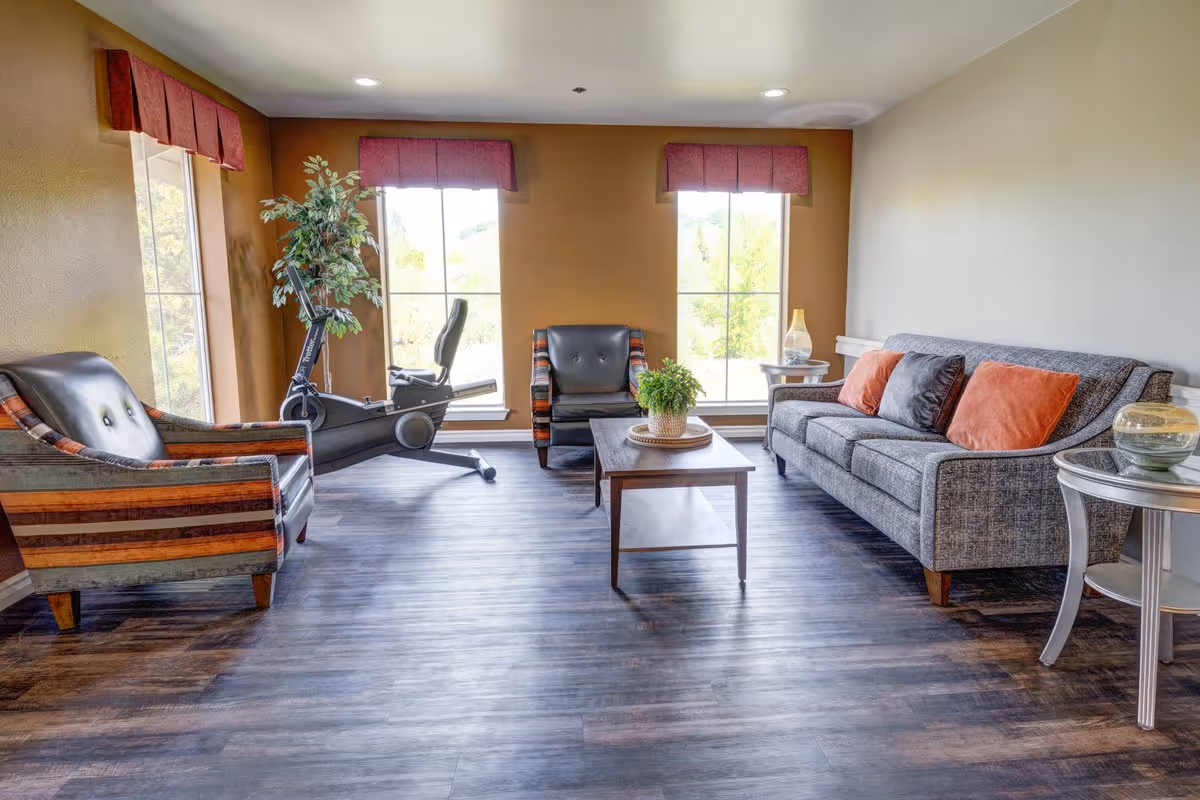 A bright living room with large windows covered by red valances, featuring a gray sofa with orange and gray pillows, two striped armchairs, a wooden coffee table with a potted plant, a side table with a lamp, and an exercise bike near the windows.