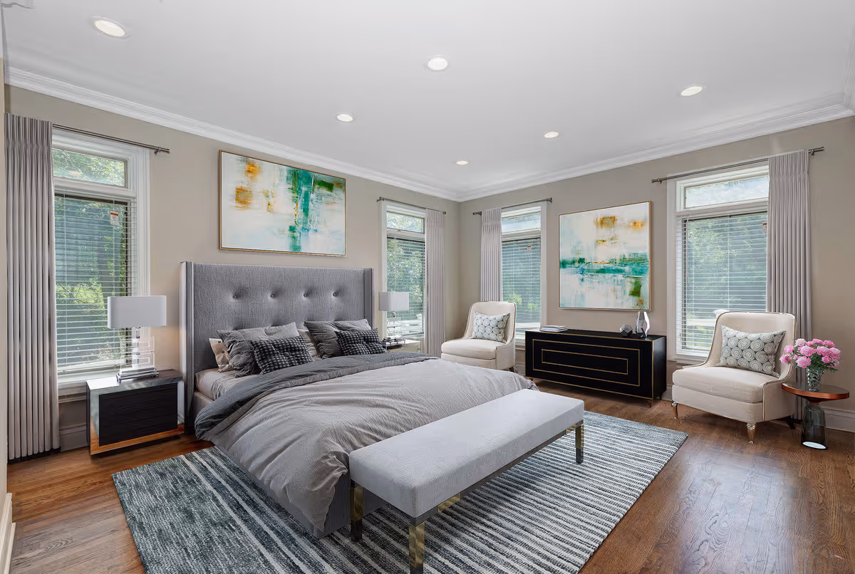 A spacious and well-lit bedroom featuring a large gray upholstered bed with multiple pillows and a gray comforter. The room has three tall windows with blinds and light gray curtains, two beige armchairs with patterned cushions, a black dresser with gold accents, and two abstract paintings on the walls. A striped area rug covers part of the wooden floor, and two modern table lamps sit on black nightstands beside the bed.