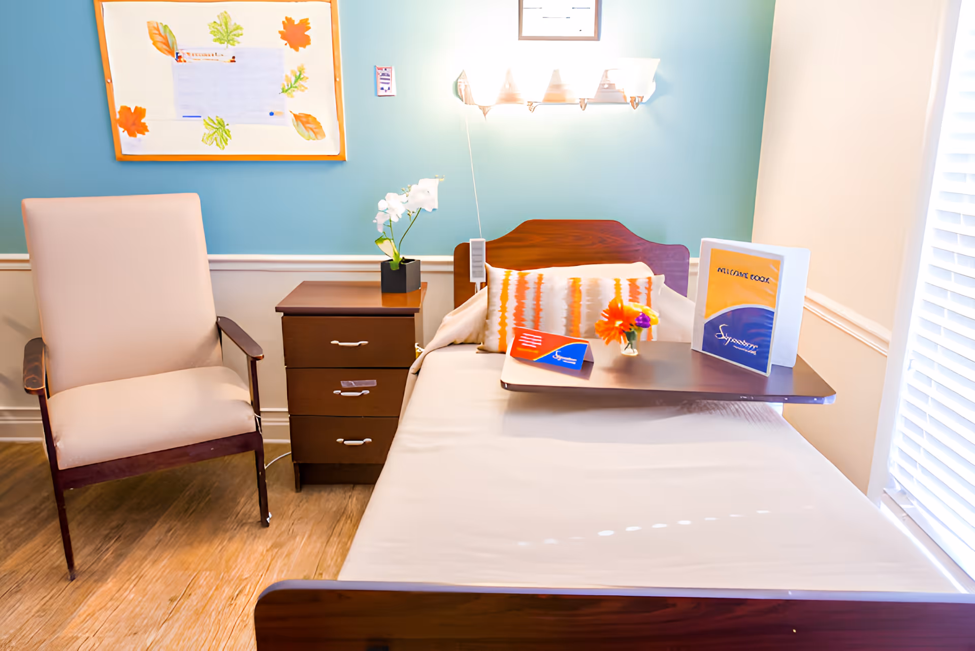 A nursing center room featuring a single bed with a wooden headboard and a tray table holding a small flower vase and informational booklets. Next to the bed is a wooden nightstand with a white orchid plant and a beige armchair. The walls are painted light blue and white, with a bulletin board decorated with autumn leaves and a wall-mounted light fixture above the bed.
