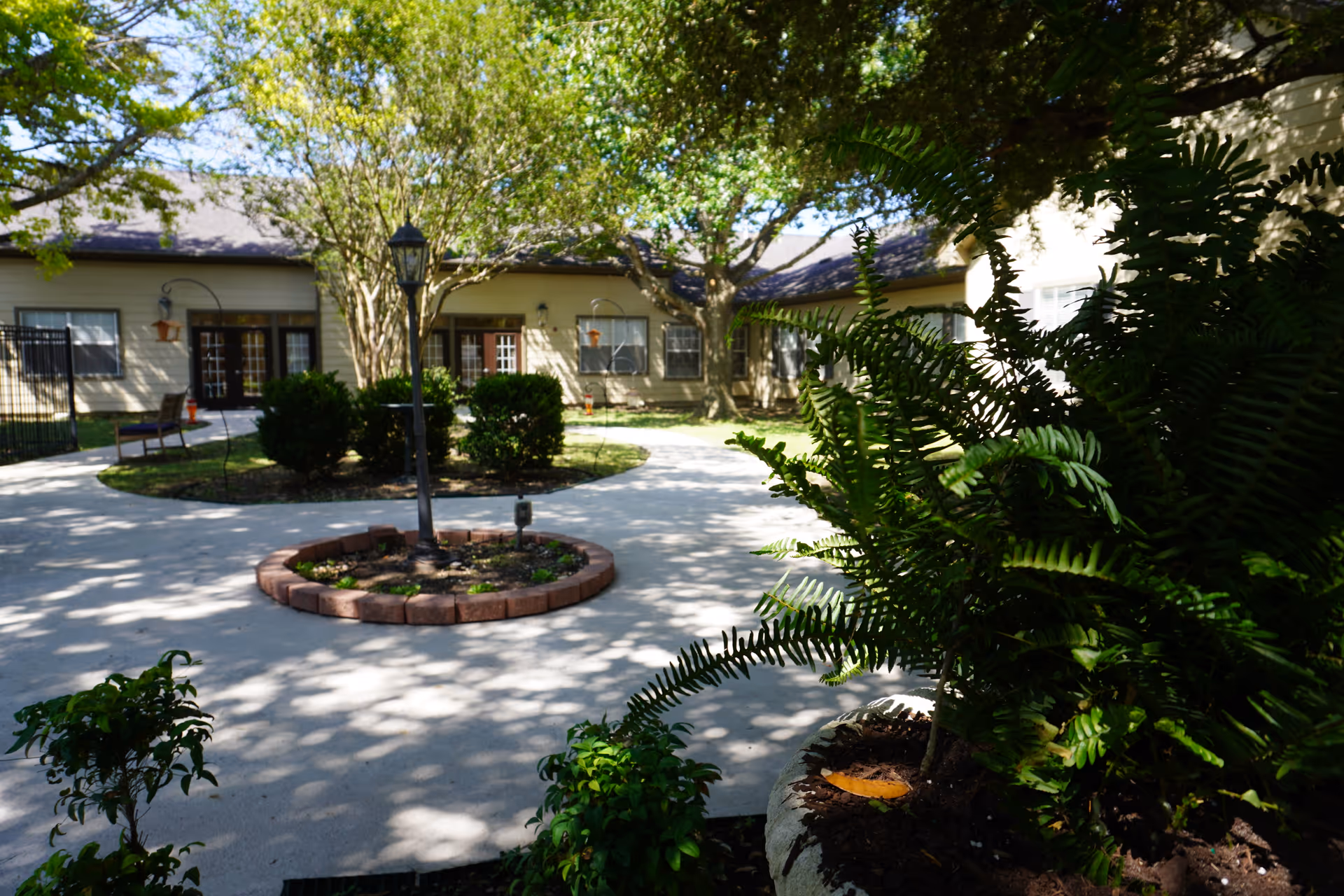 Outdoor courtyard area at Sodalis San Antonio featuring a circular paved walkway with a central garden bed bordered by bricks and a lamp post. The courtyard is surrounded by bushes, trees, and a building with windows and doors in the background. Sunlight filters through the trees, casting shadows on the ground.