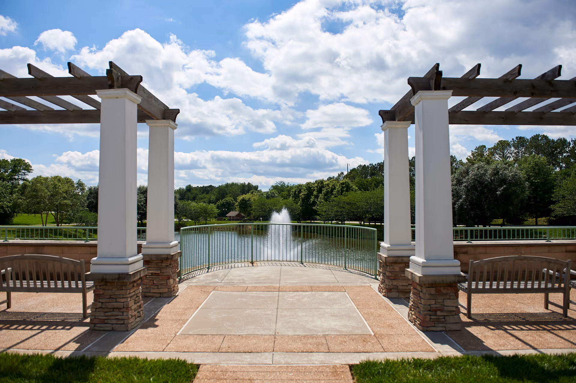 Stone-columned pergola with benches overlooking a pond and fountain under a partly cloudy sky.