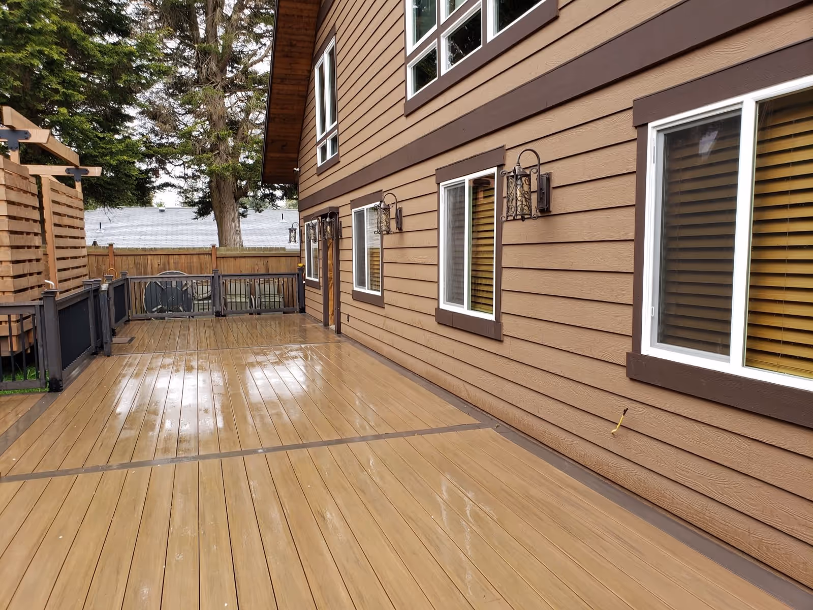 A wet wooden deck attached to a brown building with multiple windows and decorative wall-mounted lanterns. The deck is bordered by a railing and there are trees and a wooden fence in the background.