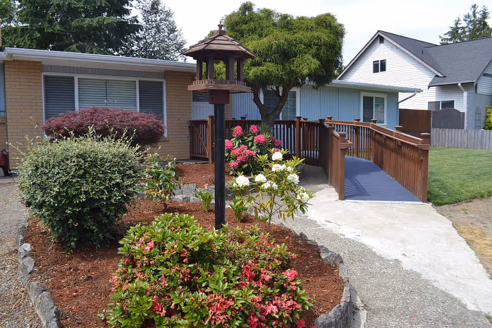 Front yard of a senior care home with a garden featuring various bushes and flowering plants, a bird feeder on a pole, a wooden ramp with railings leading to the entrance of the building, and neighboring houses in the background.
