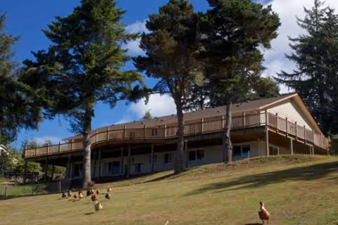 A two-story light-brown facility with a wraparound deck sits on a grassy hillside amid trees.