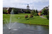 A senior living facility named Springhill Senior Living with a large green lawn, a pond with a water fountain, and a building with a cupola in the background under a partly cloudy sky.