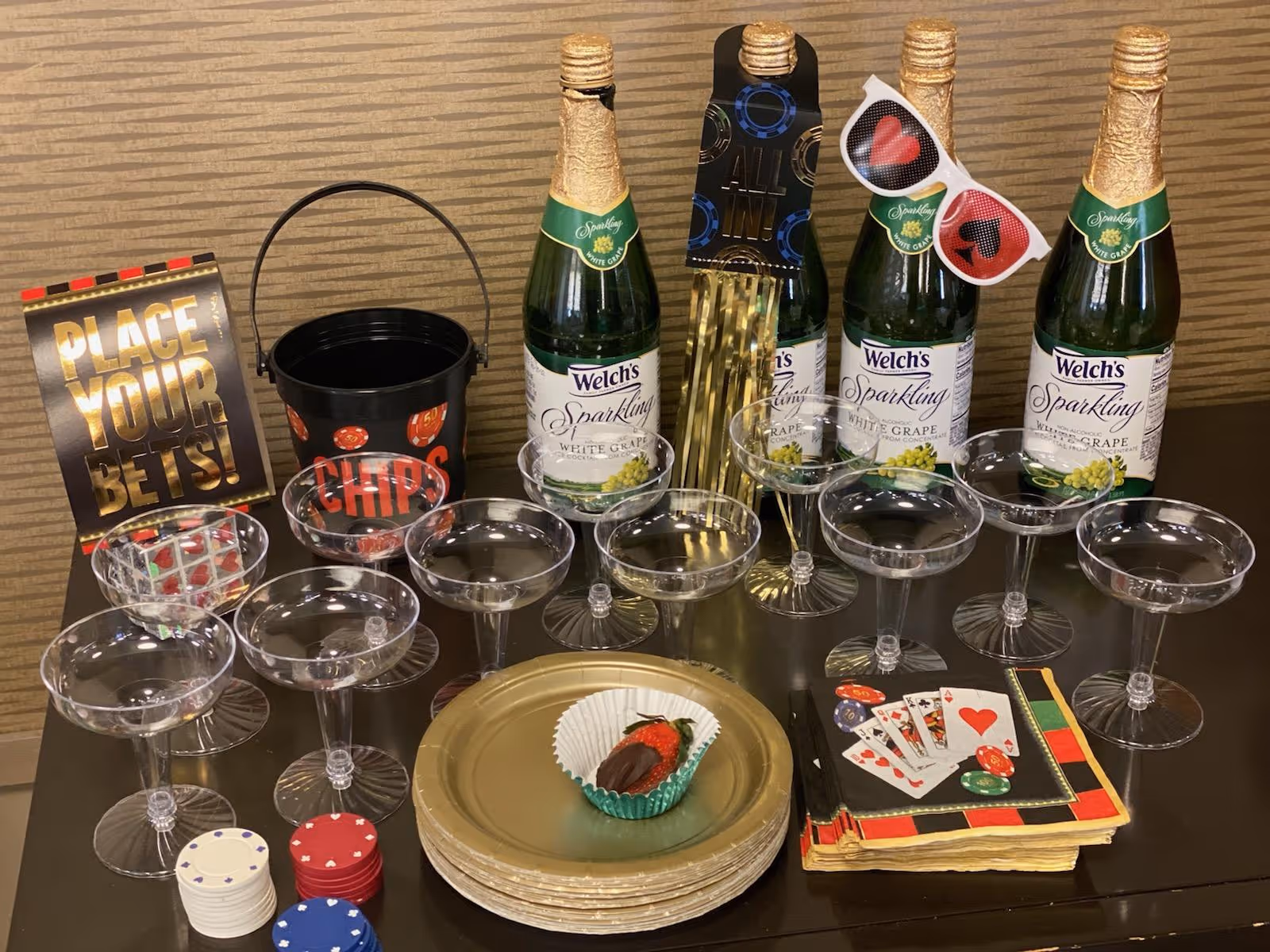 Party table with bottles of Welch's sparkling white grape, plastic champagne glasses, poker-themed decorations, chips, playing cards and a chocolate-covered strawberry.