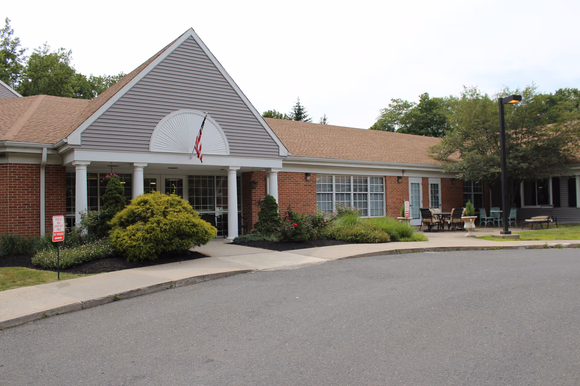 Front entrance of a single-story brick senior living facility with white columns, an American flag, landscaping and outdoor seating.