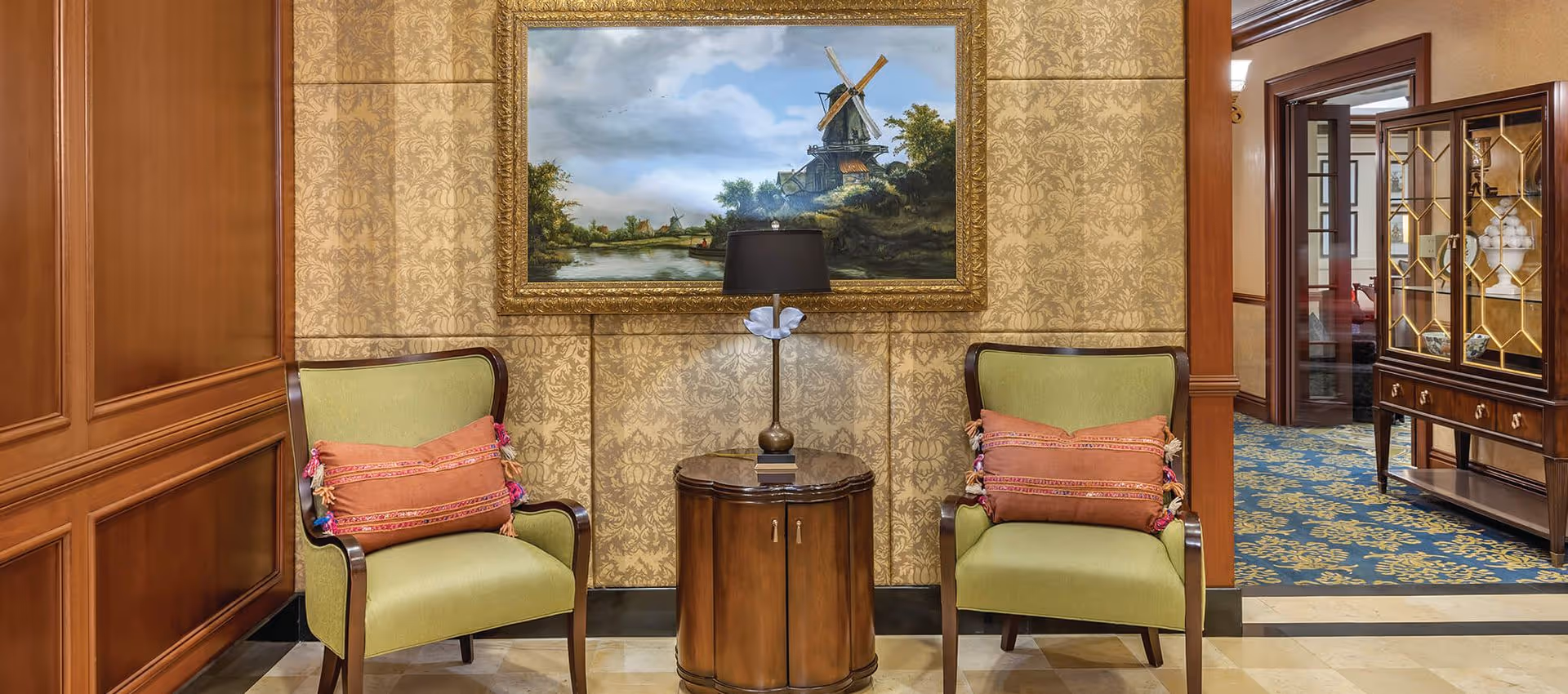 Two green upholstered armchairs with orange decorative pillows are positioned on either side of a small round wooden table with a lamp on it. Behind the table is a large framed painting of a windmill and a river, set against a wall with gold patterned wallpaper. To the right, there is a wooden cabinet with glass doors and a glimpse into another room with blue and gold patterned carpet.