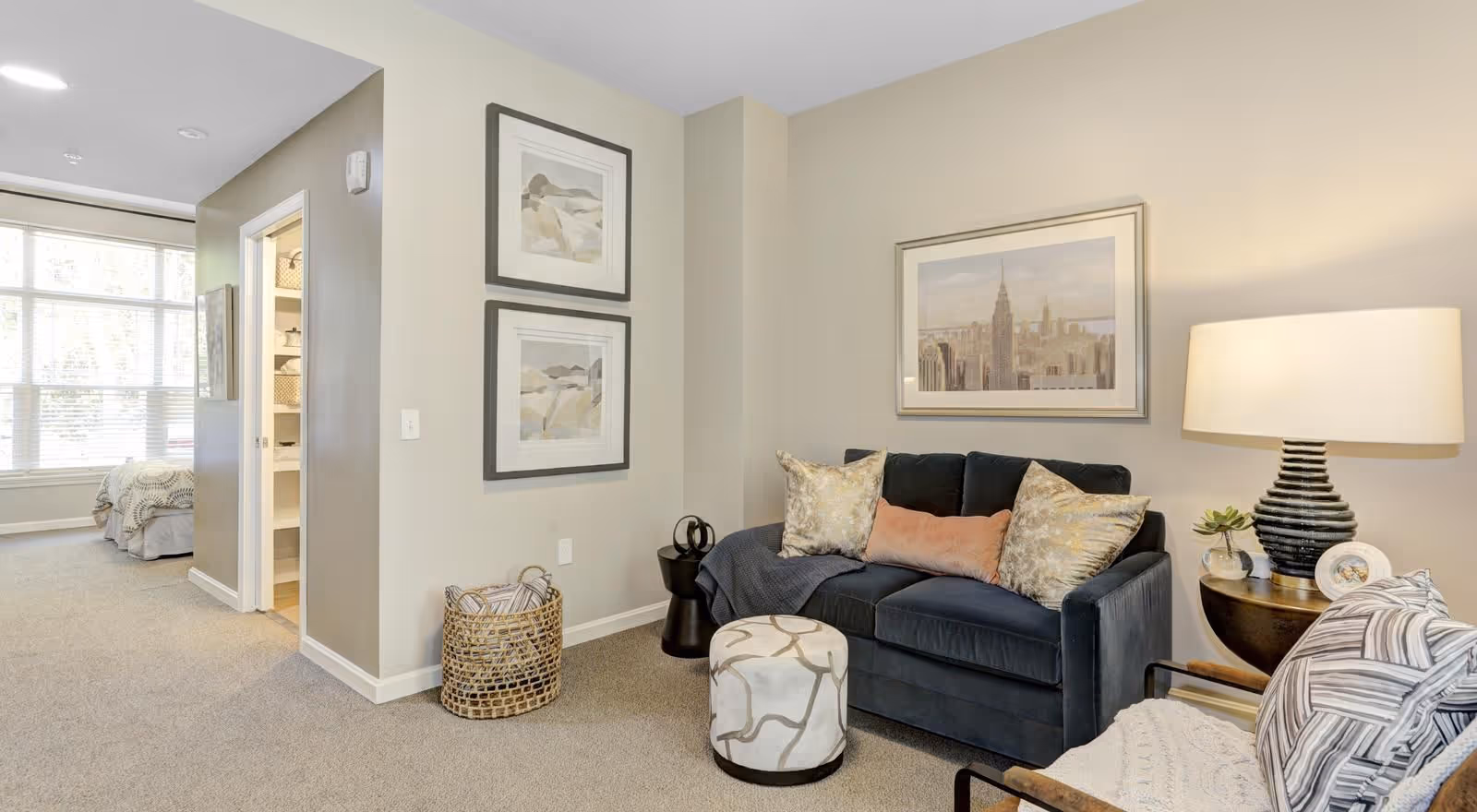 A cozy living room area in a senior living facility with a dark blue sofa adorned with decorative pillows, a patterned ottoman, a side table with a lamp and small plant, and framed artwork on the walls. In the background, a bedroom with a large window and bed is partially visible.