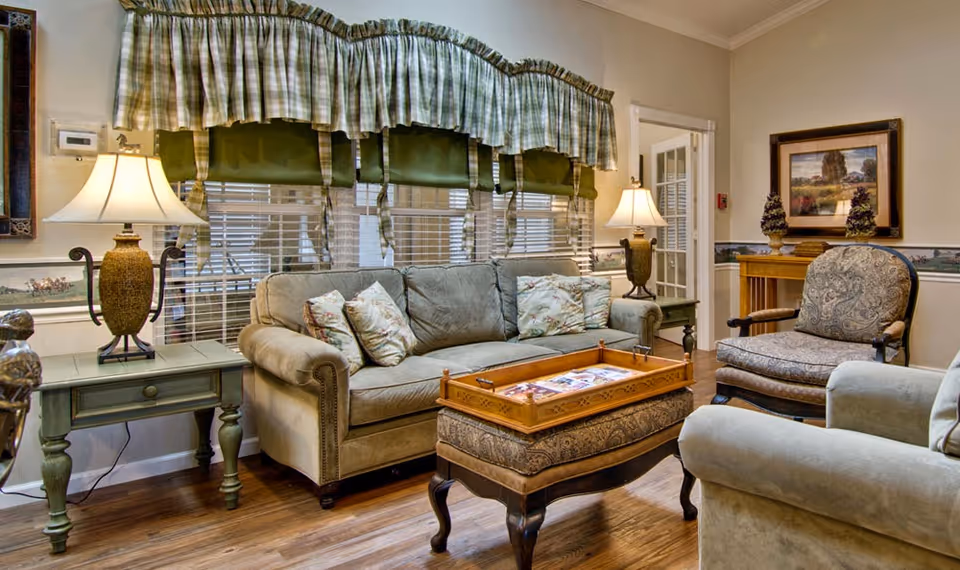 A cozy living room with a beige sofa adorned with floral pillows, a patterned armchair, and a cushioned ottoman with a wooden tray on top. Two table lamps with decorative bases are placed on side tables beside the sofa. The room features wooden flooring, a window with green and plaid valances, and a framed landscape painting on the wall.