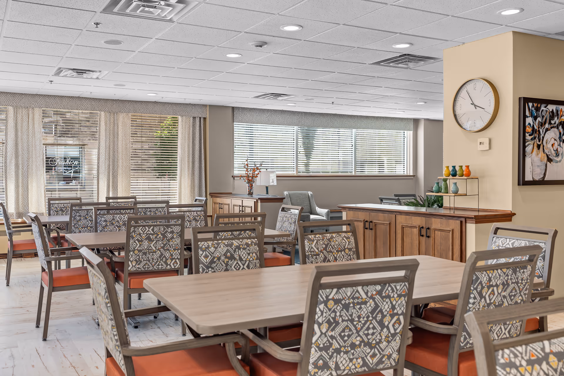 A bright dining room with multiple tables and chairs featuring patterned backs and orange cushions. Large windows with blinds allow natural light to fill the room. There is a wooden cabinet against the wall, a clock, a colorful vase display, and a framed floral painting on the wall.