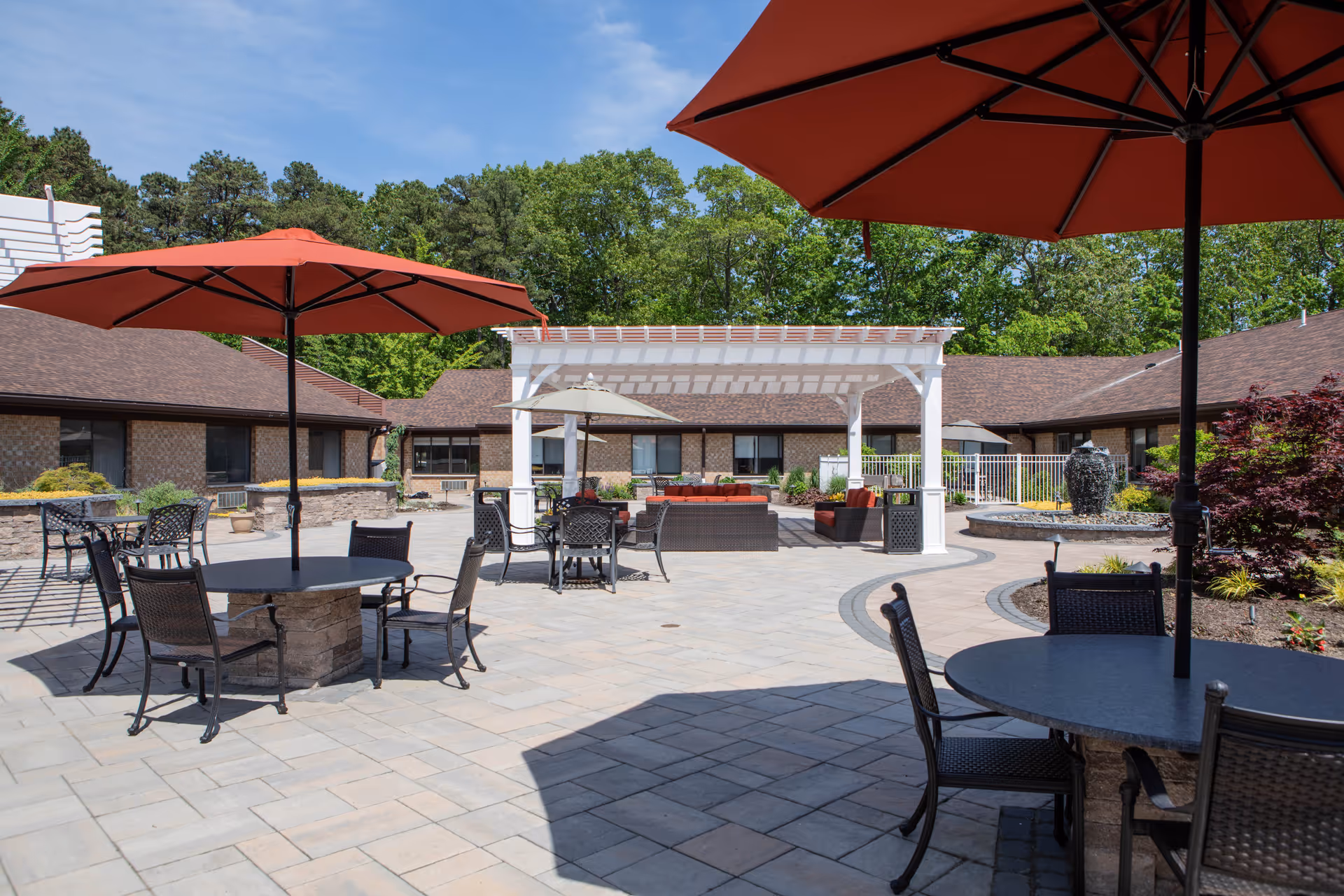 Outdoor patio area at Harmony Village at CareOne Jackson with round tables and chairs under large red umbrellas, a white pergola with seating underneath, surrounded by a single-story brick building and greenery in the background under a blue sky.