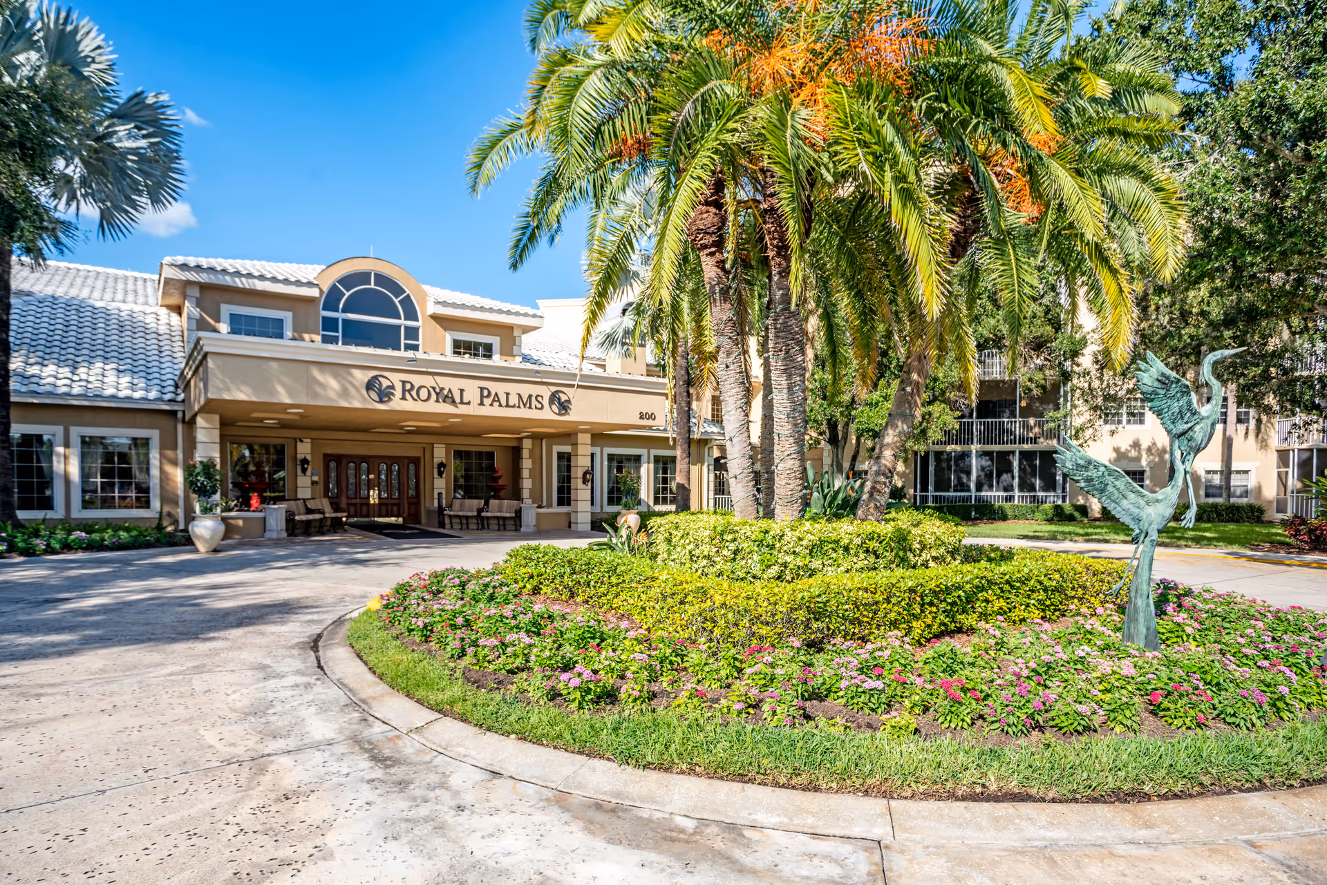 Front exterior view of the Royal Palms facility with a circular driveway, landscaped garden featuring palm trees and flowering plants, and a sculpture of two birds. The building has a beige facade with large windows and a covered entrance.