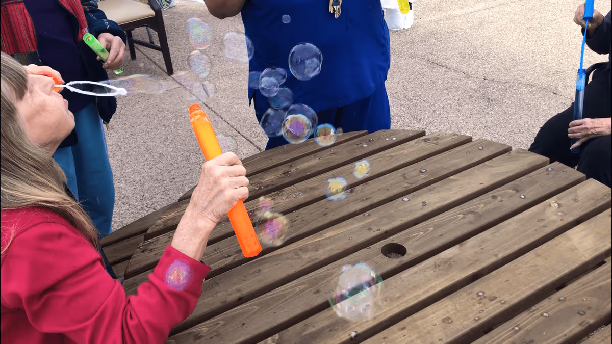 A group of elderly individuals outdoors blowing soap bubbles around a wooden picnic table on a paved surface.