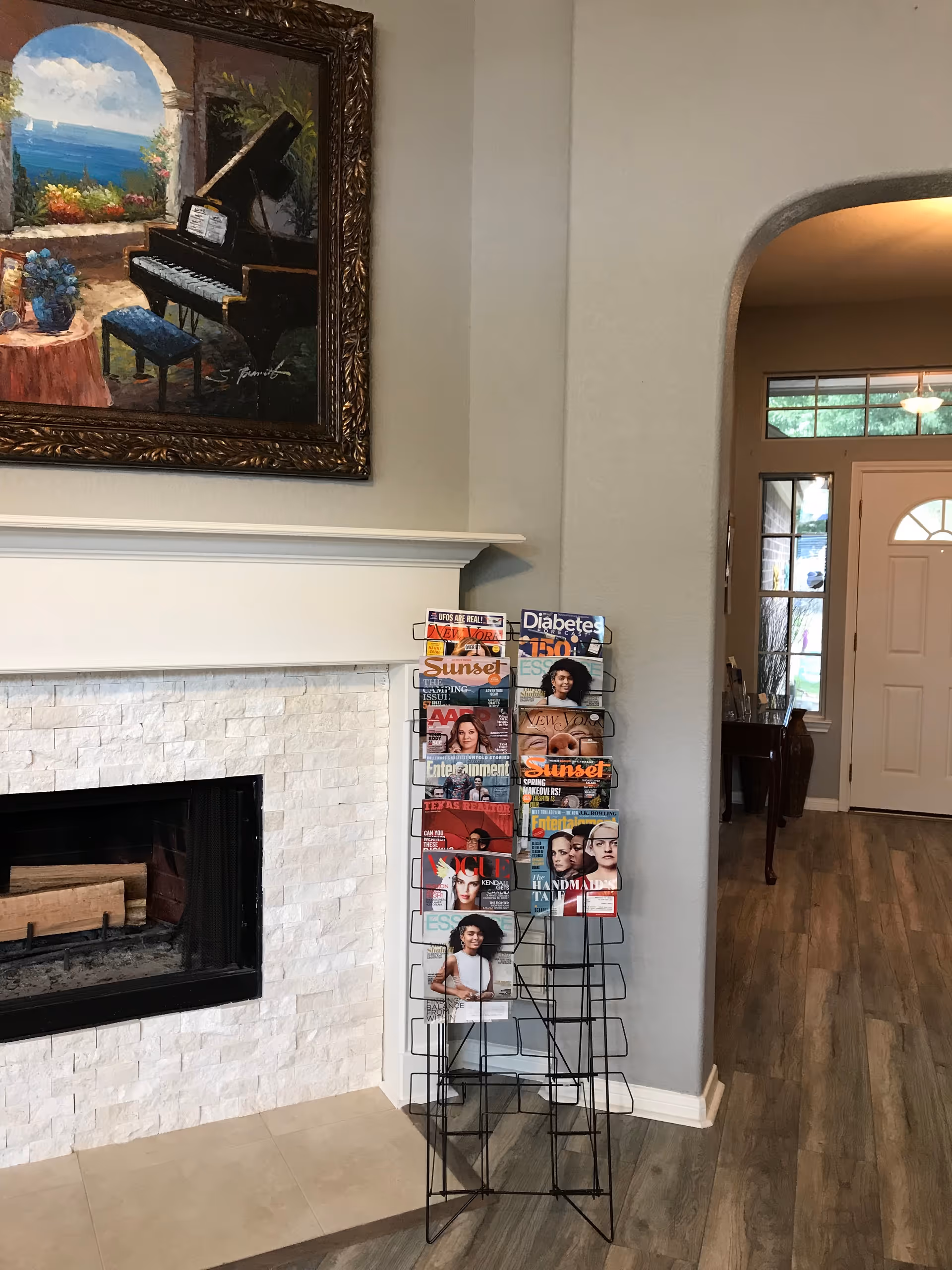 Interior view of a senior care home showing a white stone fireplace with a wooden mantel. Above the mantel is a framed painting of a piano by a window overlooking a seascape. Next to the fireplace is a black metal magazine rack filled with various magazines. The room has light-colored walls and wood flooring, with an arched doorway leading to an entrance door with windows.