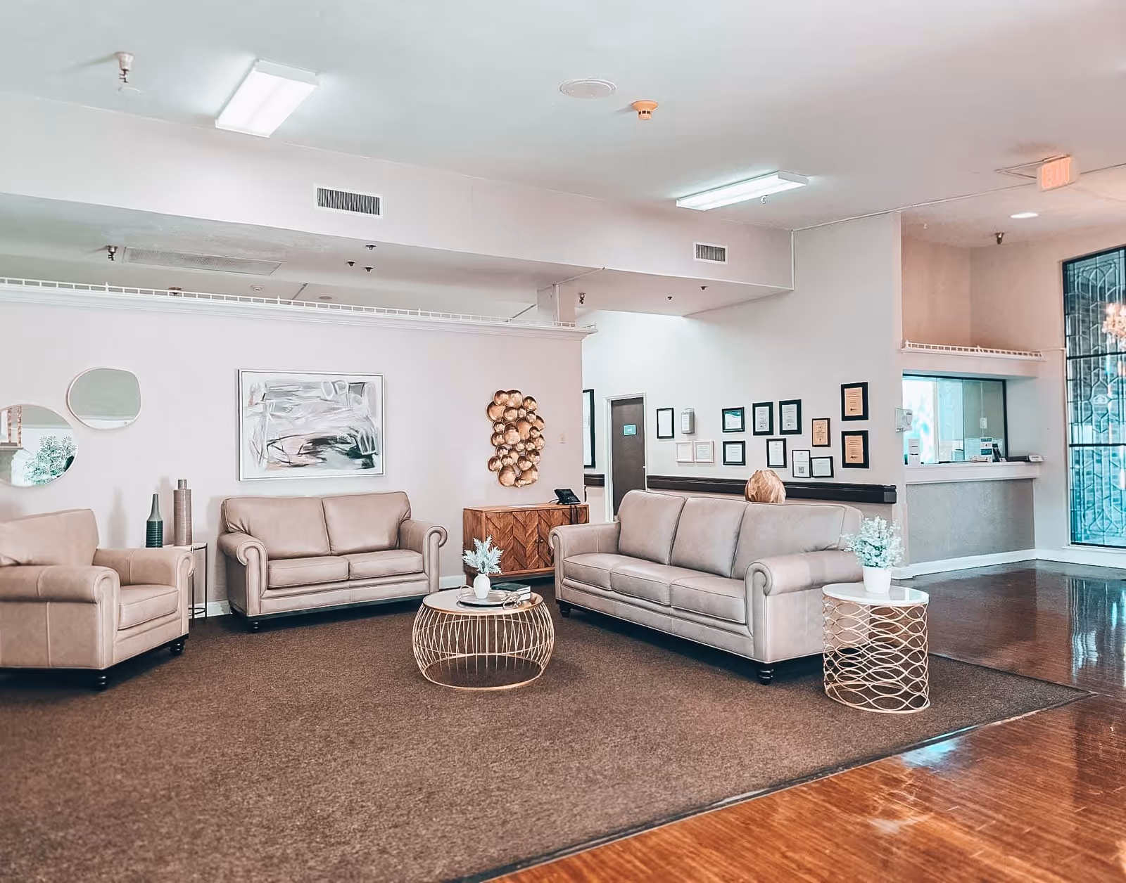 A spacious and well-lit living room area with beige leather sofas and armchairs arranged around two round metal coffee tables with decorative plants. The walls are decorated with mirrors, abstract art, and a wall sculpture. There is a reception desk visible in the background with framed certificates on the wall and a large window with decorative glass panels.