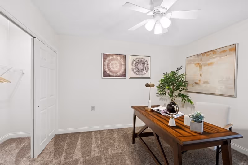 A small office room with beige carpet and white walls. There is a wooden desk with a lamp, a globe, some books, and small potted plants on it. Behind the desk is a white upholstered chair. On the wall above the chair are three framed abstract art pieces. A ceiling fan with lights is mounted on the ceiling. To the left, there is a closet with white sliding doors and a hanging rod with a few hangers.