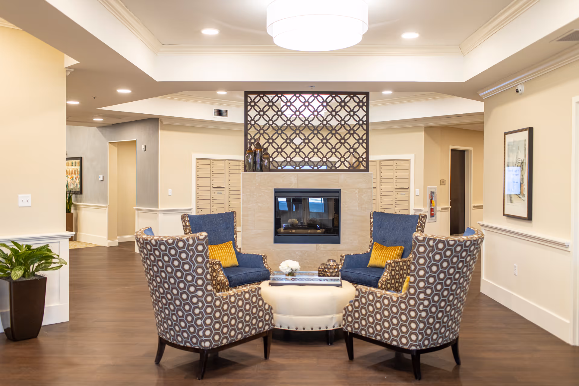 A cozy senior living lobby with patterned armchairs arranged around an ottoman in front of a central fireplace and decorative screen.