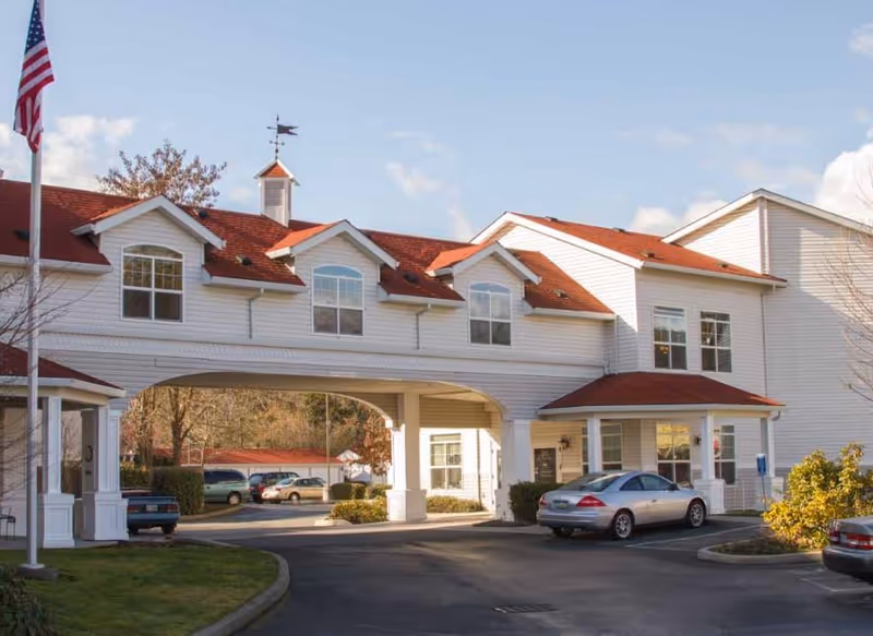 White multi-story retirement facility with red roofs, a covered entrance, and parked cars.