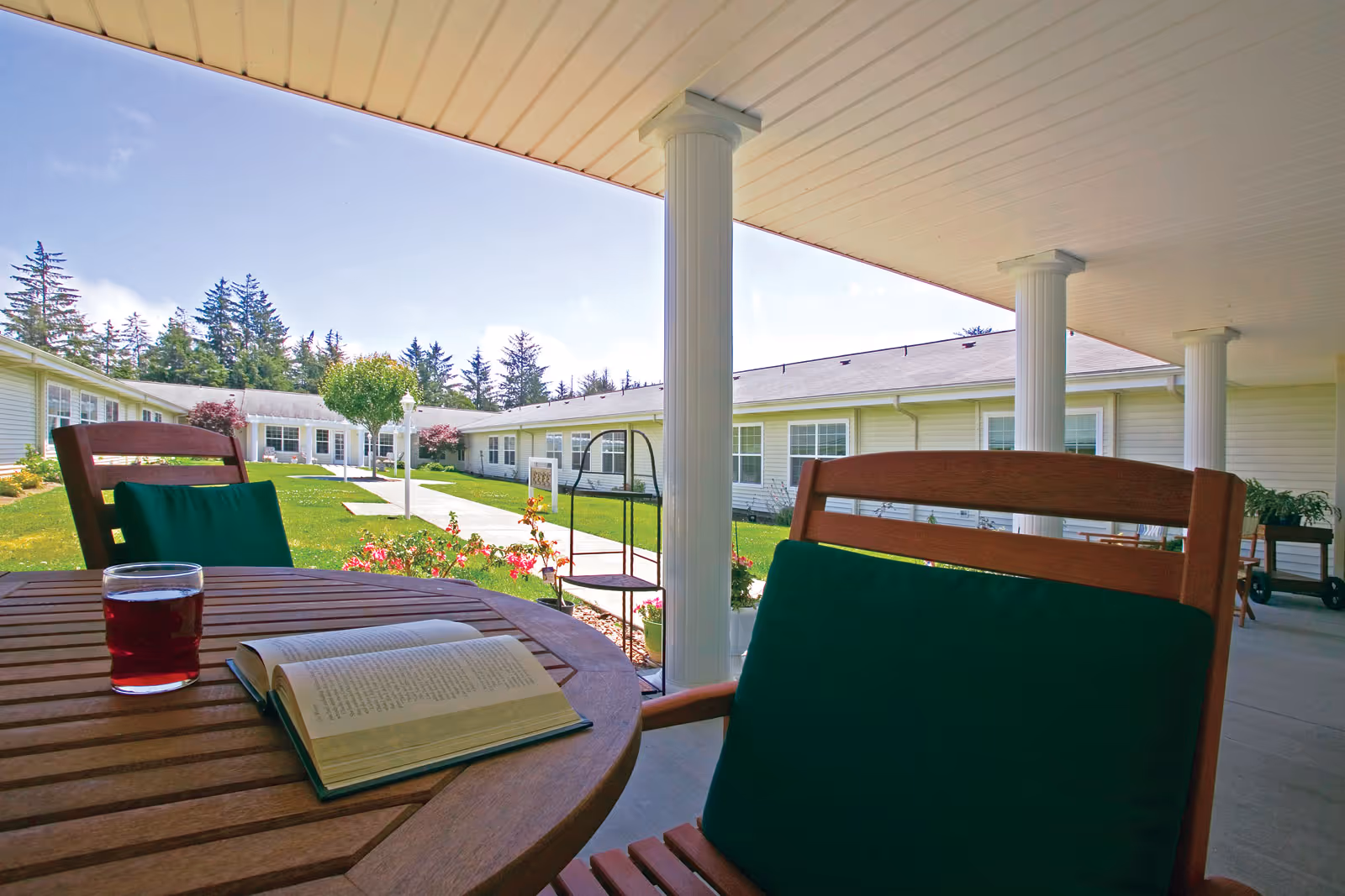 View from a covered patio area with wooden chairs and a round wooden table holding an open book and a glass of red drink. The patio overlooks a green lawn with flowers, a walkway, and a single-story building with white siding and multiple windows. Trees are visible in the background under a clear blue sky.