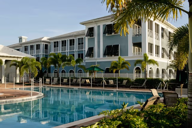 Outdoor view of a senior living facility named Heron Point Independent Living of Vero Beach, featuring a large swimming pool surrounded by lounge chairs, palm trees, and a multi-story building with balconies and windows in the background under a clear sky.