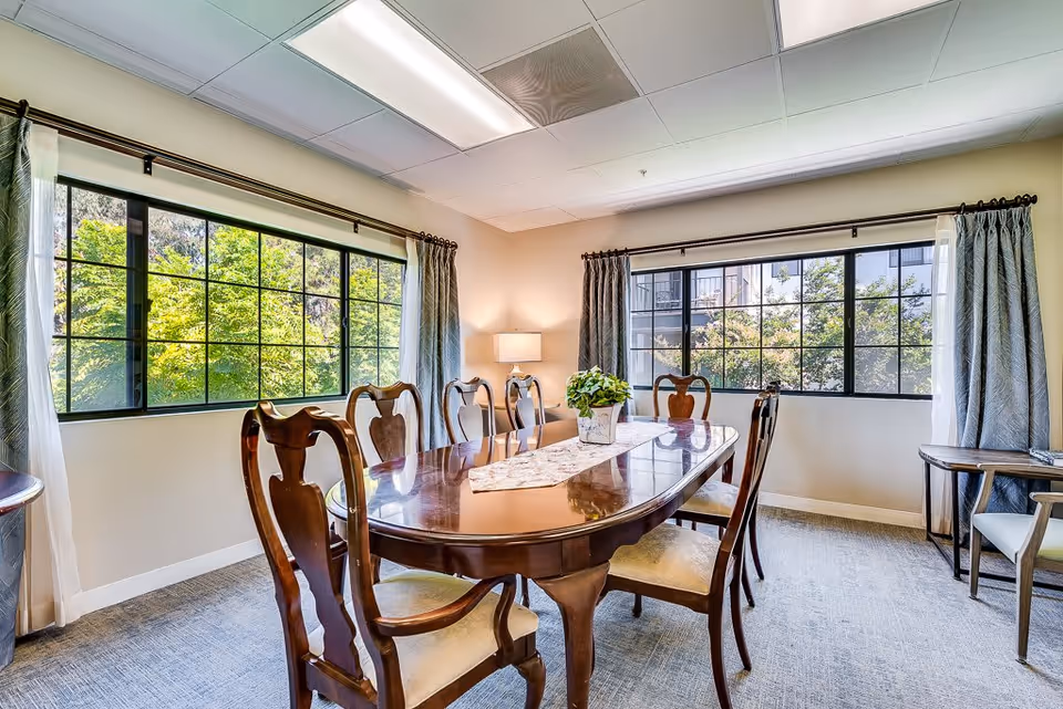 A bright dining room with a polished wooden dining table surrounded by six matching chairs. The room has large windows with black frames and curtains, allowing natural light to fill the space. A small potted plant and a decorative runner are placed on the table. The walls are light-colored, and a lamp is visible in the corner.