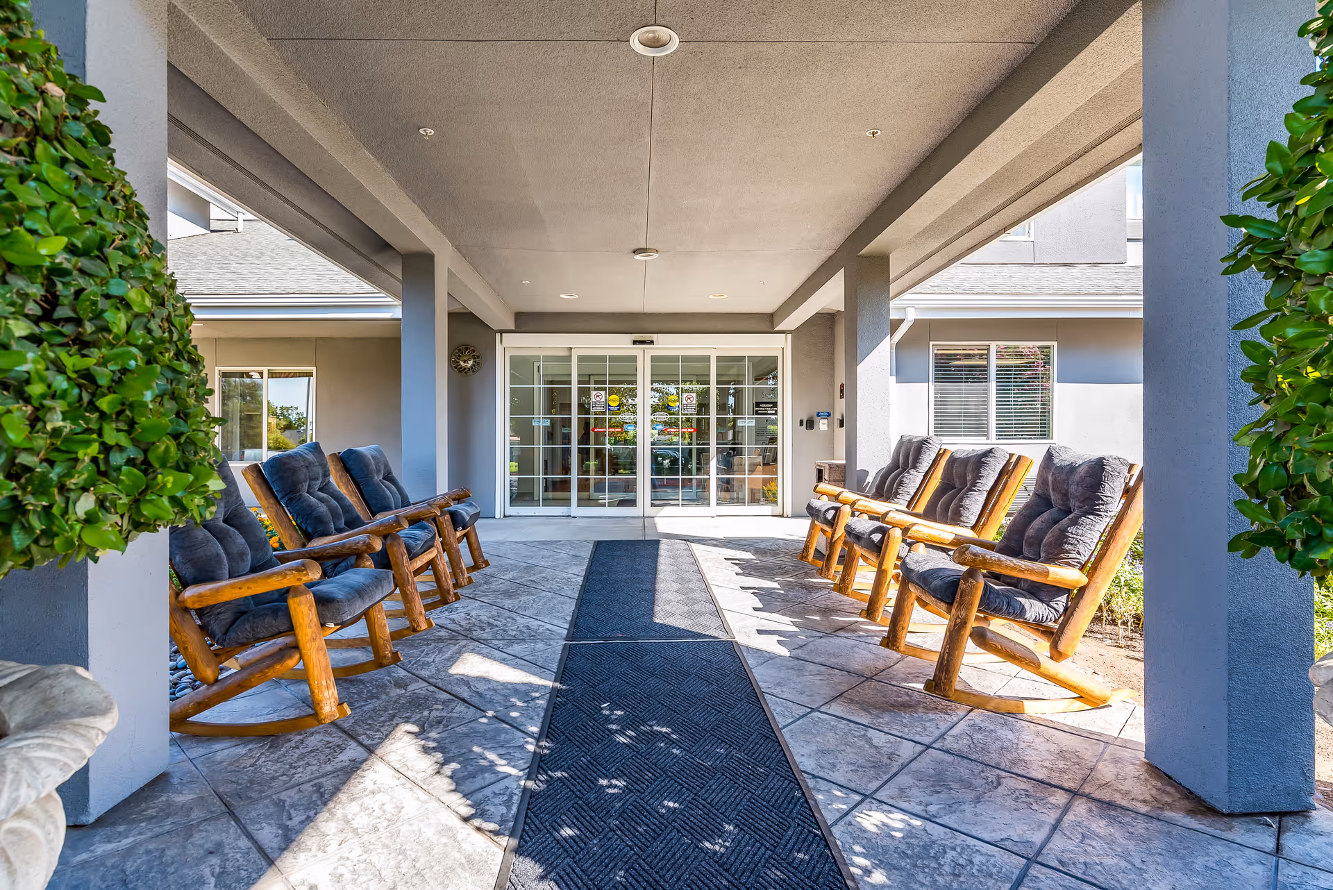 Covered entrance to a senior living facility with sliding glass doors and rows of cushioned wooden rocking chairs on either side of the walkway.