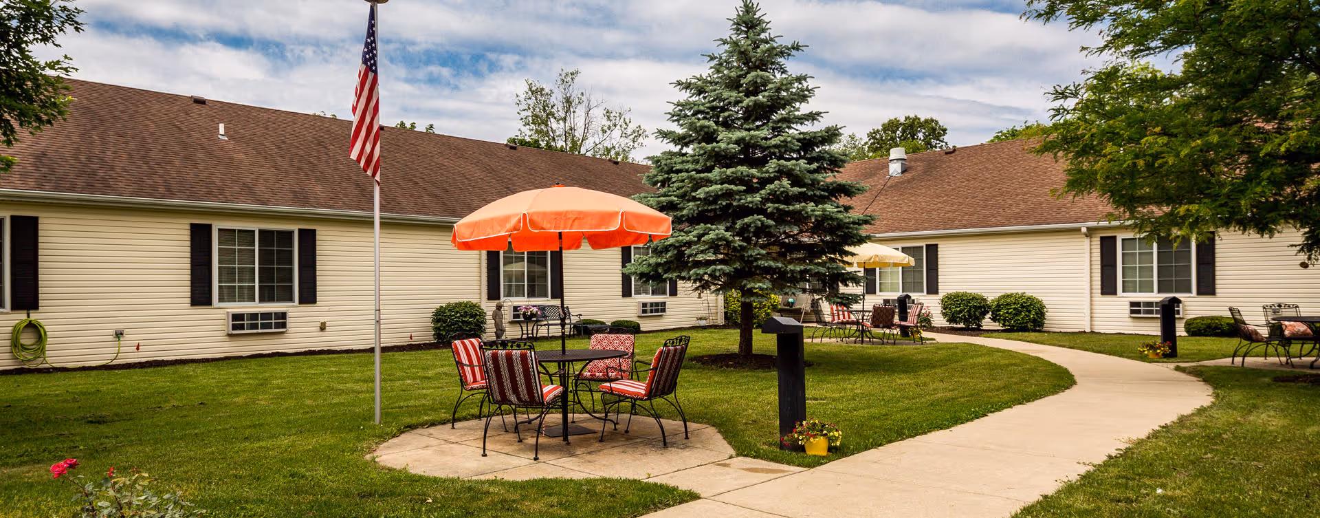 Outdoor courtyard area at Bickford of Battle Creek with a circular concrete patio featuring a table and chairs under an orange umbrella, surrounded by green grass, trees, and a beige building with brown roof and windows in the background.
