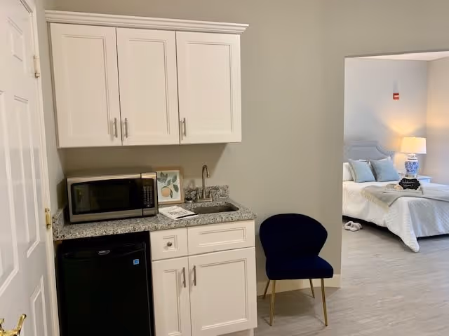 A small kitchenette area with white cabinets, a granite countertop, a microwave, a small sink, and a mini refrigerator. Next to the kitchenette is a dark blue chair with gold legs. In the background, there is a bedroom with a bed, pillows, a bedside table with a lamp, and slippers on the floor.