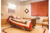 A neatly arranged nursing home bedroom with a single bed made up with pillows and blankets. The room has beige walls with patterned wallpaper, a window with closed wooden blinds, a small air conditioning unit below the window, a chair, and framed artwork on the walls.