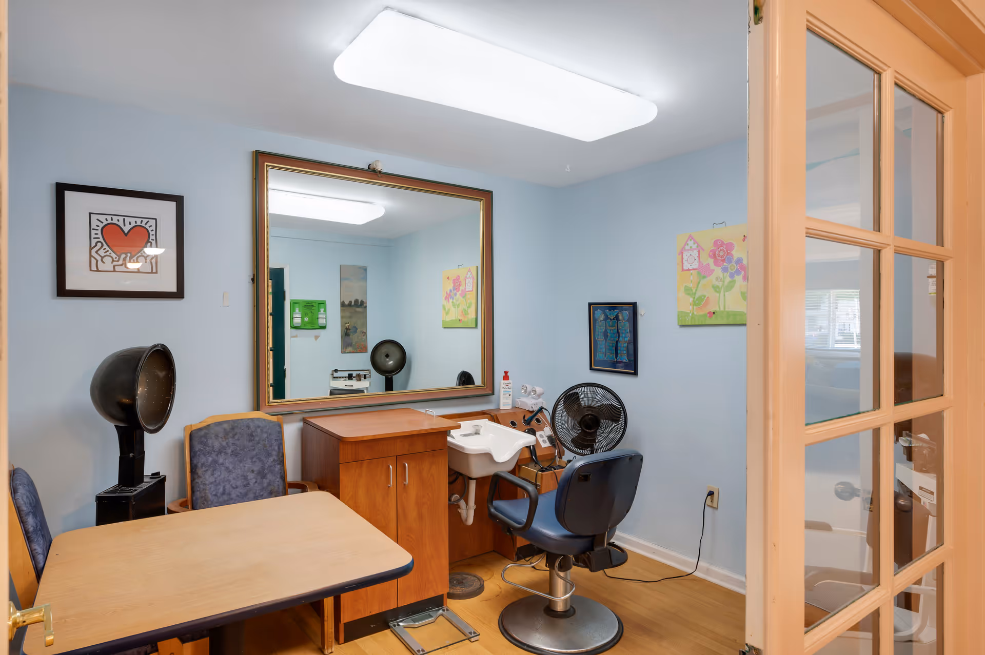 Small room with light blue walls featuring a large mirror, a hair washing station with a blue salon chair, a black hair dryer, a wooden table with two blue chairs, and colorful artwork on the walls. The room has a wooden floor and a glass-paneled door partially open.