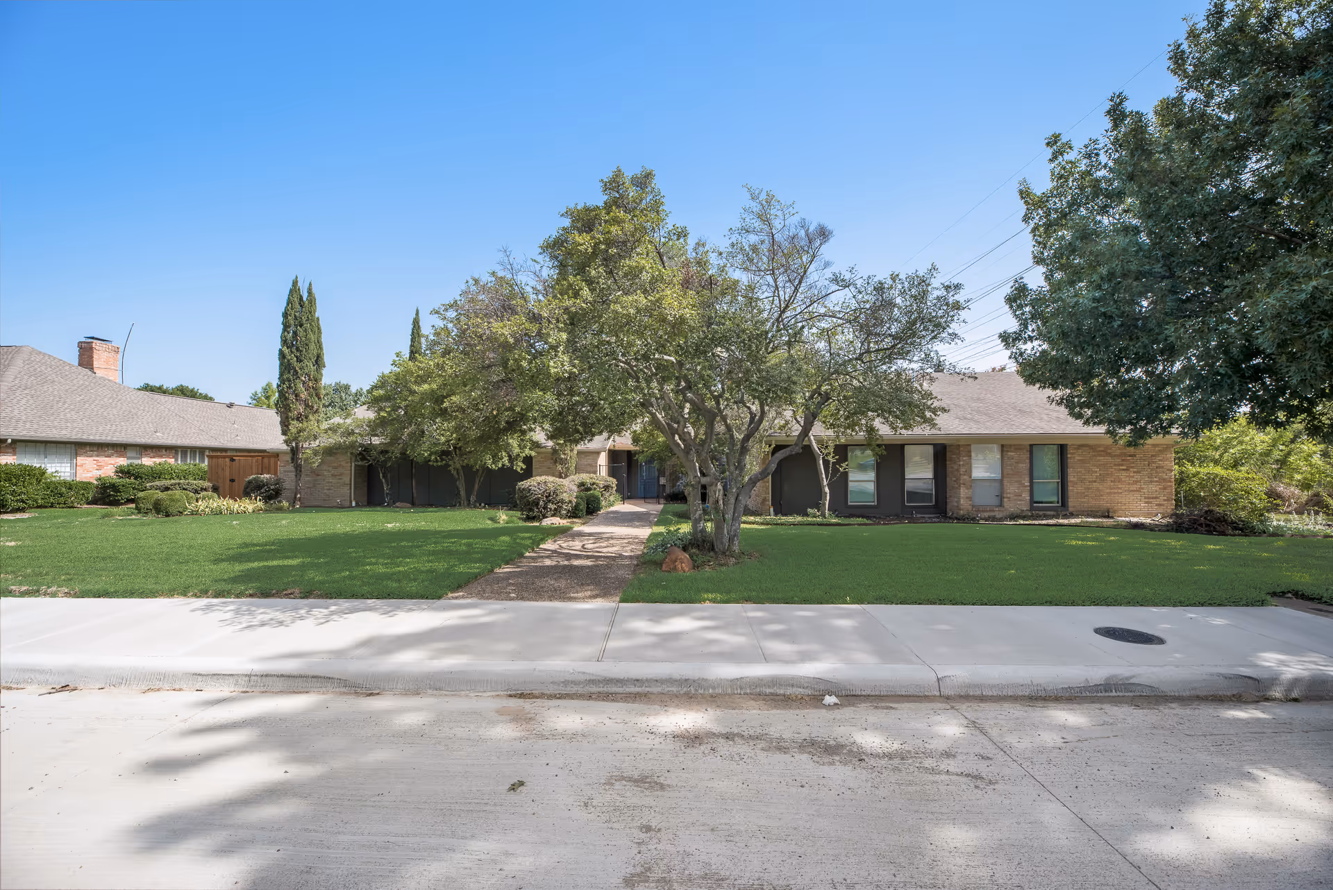 Single-story brick building with a well-maintained lawn, trees, and a sidewalk in front under a clear blue sky.
