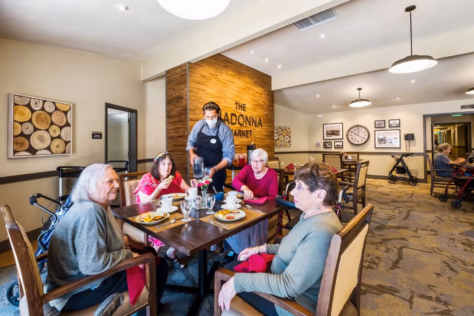 Several elderly residents seated around a dining table in a bright assisted-living dining room as a staff member pours coffee.