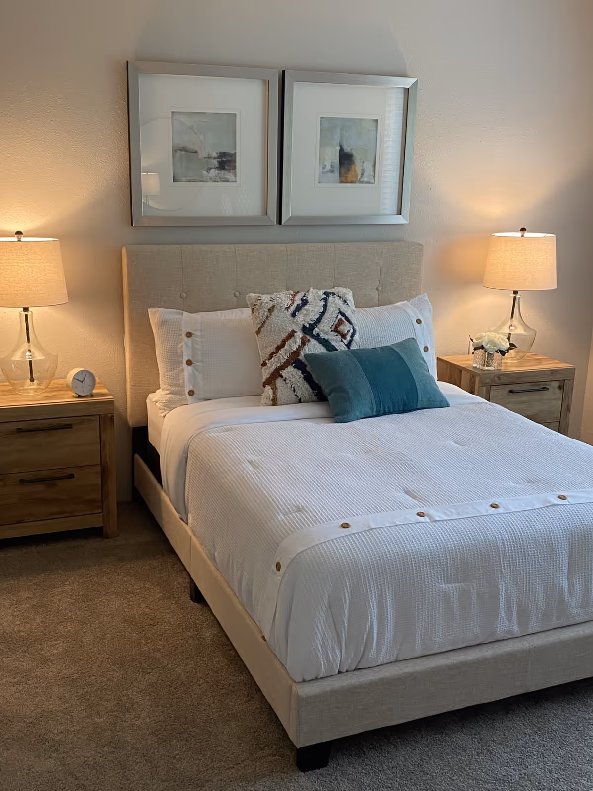 A neatly made bed with a beige upholstered headboard, white bedding with button details, and three decorative pillows in white, teal, and patterned colors. On each side of the bed are wooden nightstands with glass lamps emitting warm light. Above the bed are two framed abstract paintings. The room has beige carpet and light-colored walls.