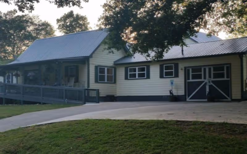 Exterior view of a single-story building with light yellow siding and a metal roof, surrounded by trees and a grassy lawn. The building features several windows, a ramp leading to a porch area, and a garage door on the right side.