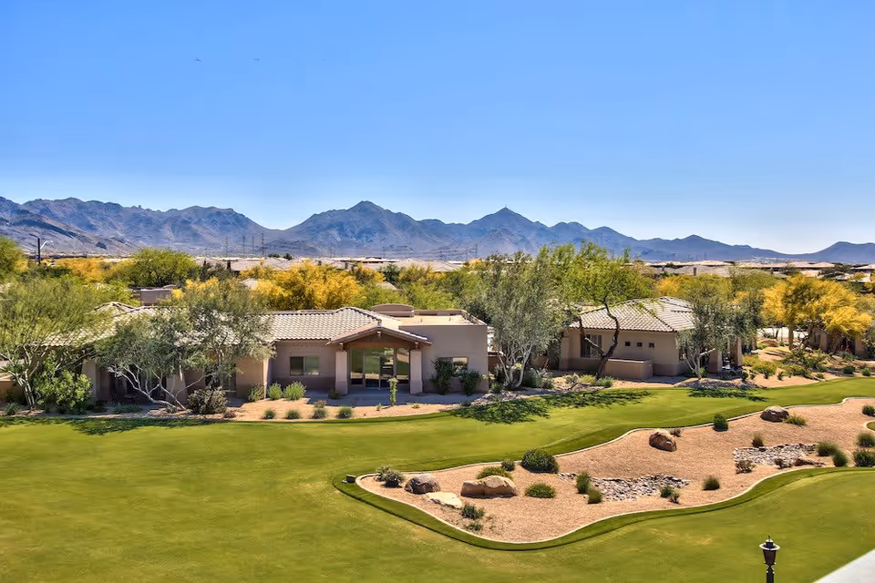 View of a senior living facility with single-story buildings surrounded by desert landscaping and green lawns, set against a backdrop of mountains under a clear blue sky.