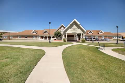 Front exterior view of a single-story senior living facility building with a peaked entrance canopy, surrounded by well-maintained green lawns and intersecting concrete walkways under a clear blue sky.