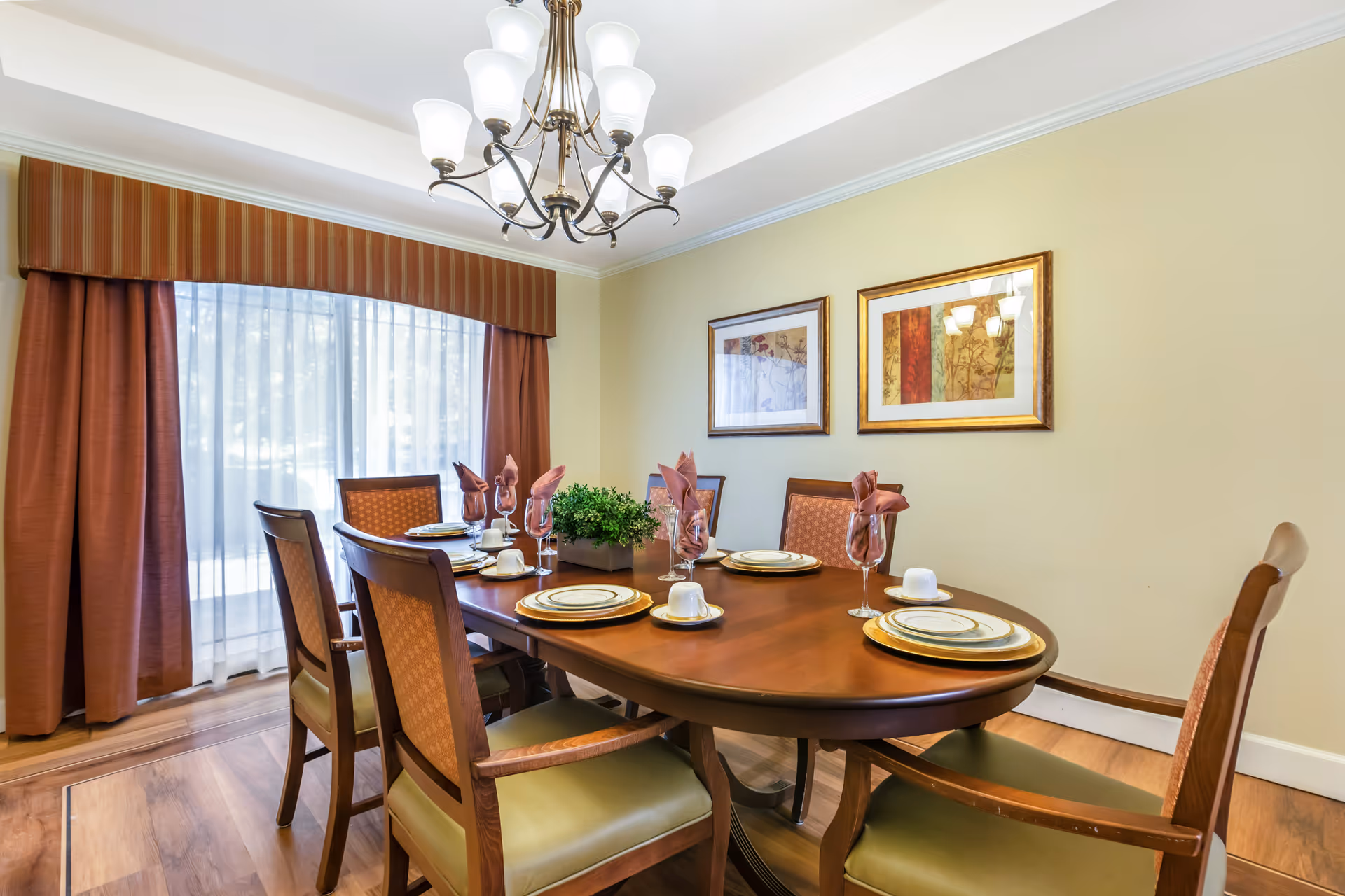 A dining room with a wooden oval table set for six people. Each place setting includes a plate, cup, and a wine glass with a folded napkin. The room has wooden chairs with cushioned seats, a large window with sheer white curtains and rust-colored drapes, two framed artworks on a beige wall, and a chandelier with multiple lights hanging from the ceiling.