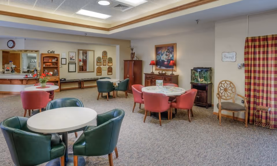A cozy common area in a senior living facility featuring several round tables with chairs arranged around them. The room has a carpeted floor, a small fish tank on a wooden stand, a cabinet with decorative items and lamps, and a wall painting. There is a small café counter in the background with a clock above it and a sign that reads 'Café'. The walls are decorated with plaques, mirrors, and a sign that says 'Sweet'. A window with red and yellow checkered curtains is visible on the right side.