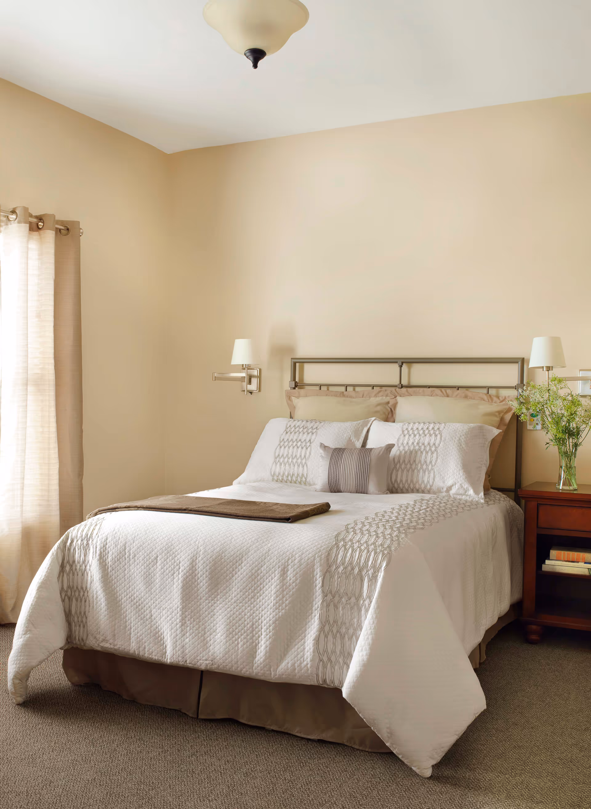 A neatly made bed with white and beige bedding in a bedroom with beige walls. There are two wall-mounted lamps on either side of the bed, a wooden nightstand with books and a vase of flowers, and beige curtains covering a window.