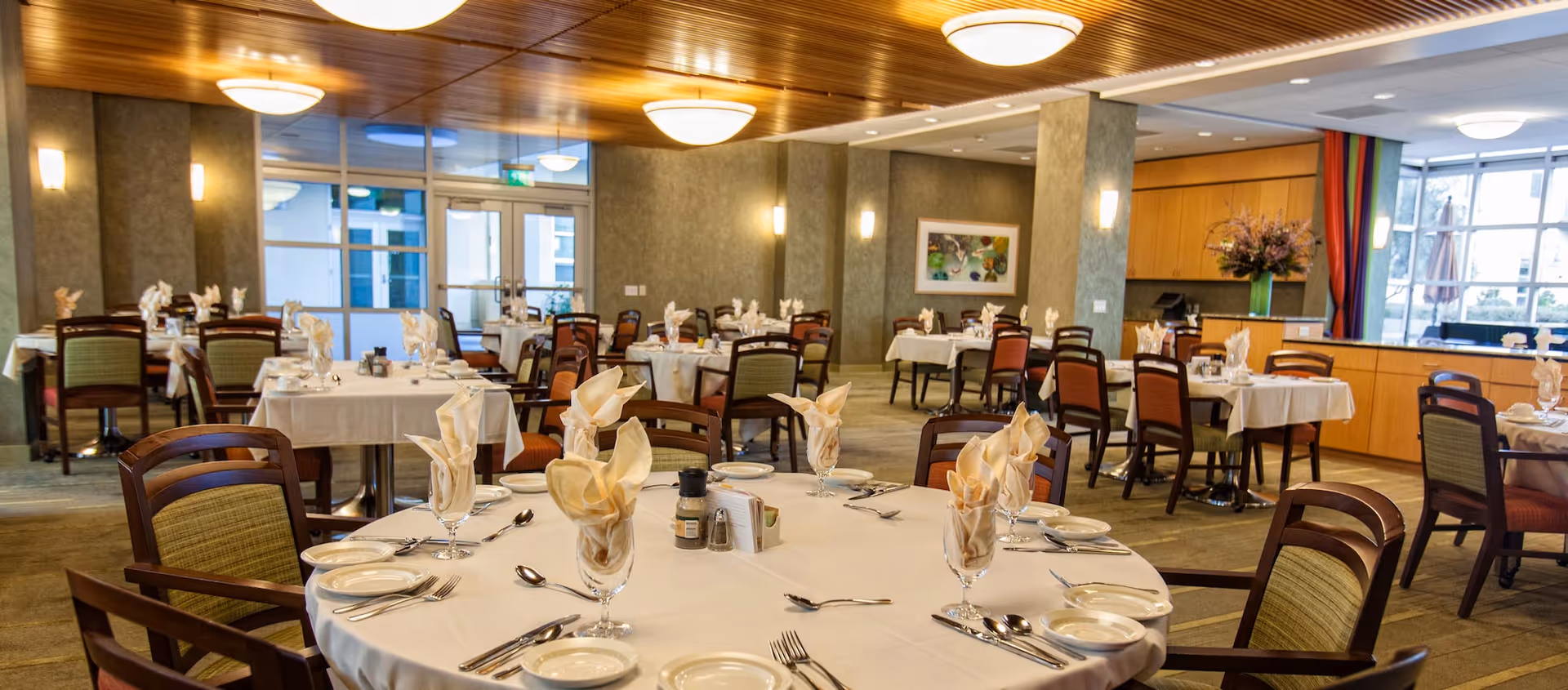 A spacious dining room with multiple round and rectangular tables set with white tablecloths, folded napkins in glasses, plates, and silverware. The room features wooden ceiling panels, soft lighting fixtures, and large windows allowing natural light to enter. Chairs with wooden frames and cushioned seats surround the tables.