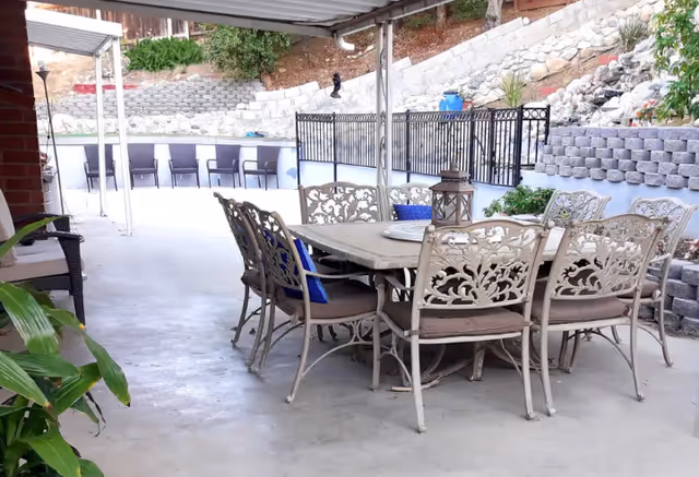 Outdoor covered patio area with a large rectangular metal table surrounded by eight ornate metal chairs with cushions. There are blue pillows on some chairs and a decorative lantern on the table. The patio overlooks a terraced garden with stone retaining walls and some plants.
