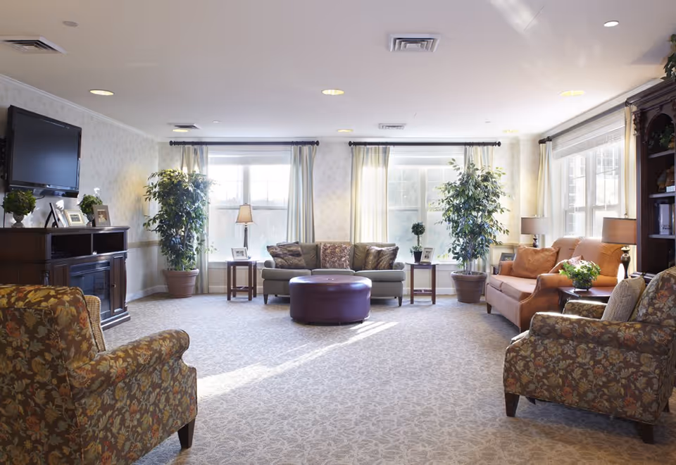 A bright and spacious living room with large windows letting in natural light. The room features a green sofa, an orange sofa, two floral armchairs, a round purple ottoman in the center, side tables with lamps and framed photos, potted plants, a wall-mounted flat-screen TV, and a wooden cabinet with decorative items.