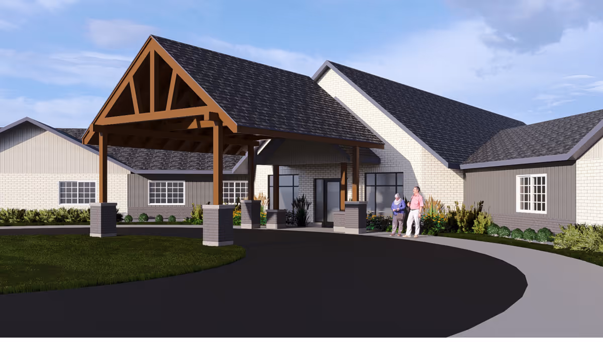 Exterior view of Boka Haven facility showing a covered entrance with wooden beams and brick pillars. Two elderly people are standing near the entrance on a curved driveway, with landscaping and shrubs around the building under a partly cloudy sky.