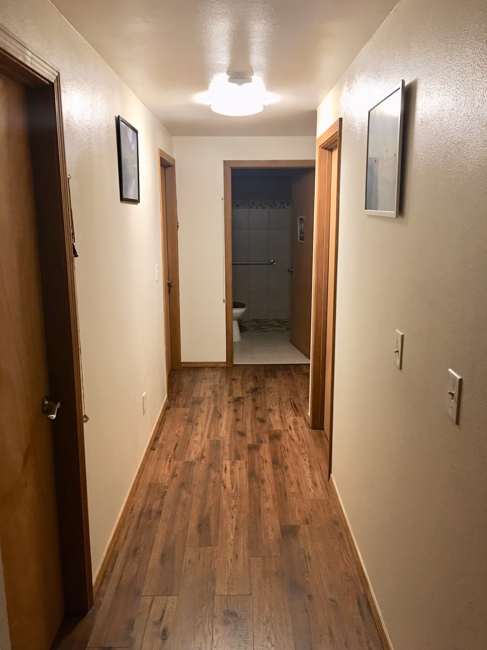 A hallway with wooden flooring and cream-colored walls leading to a bathroom at the end. There are three wooden doors along the hallway, two on the left and one on the right. The bathroom door is open, showing a toilet and tiled walls inside. Two framed pictures hang on the walls of the hallway, and a ceiling light fixture illuminates the space.