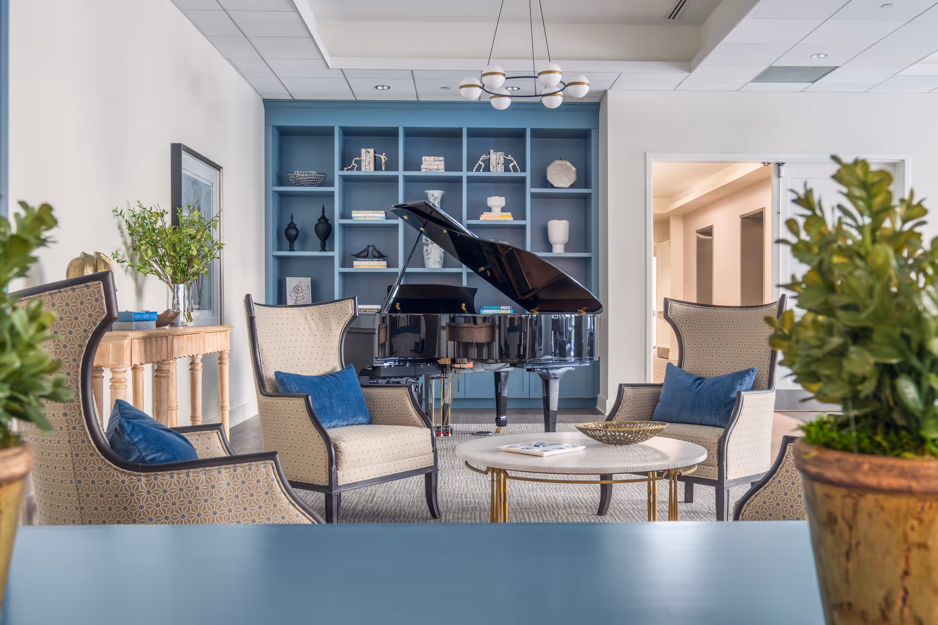 A cozy sitting area with four upholstered armchairs featuring blue cushions arranged around a round coffee table. In the background, there is a black grand piano in front of a blue built-in bookshelf filled with decorative items and books. The room has light-colored walls, a wooden console table with plants and framed artwork, and potted plants in the foreground.