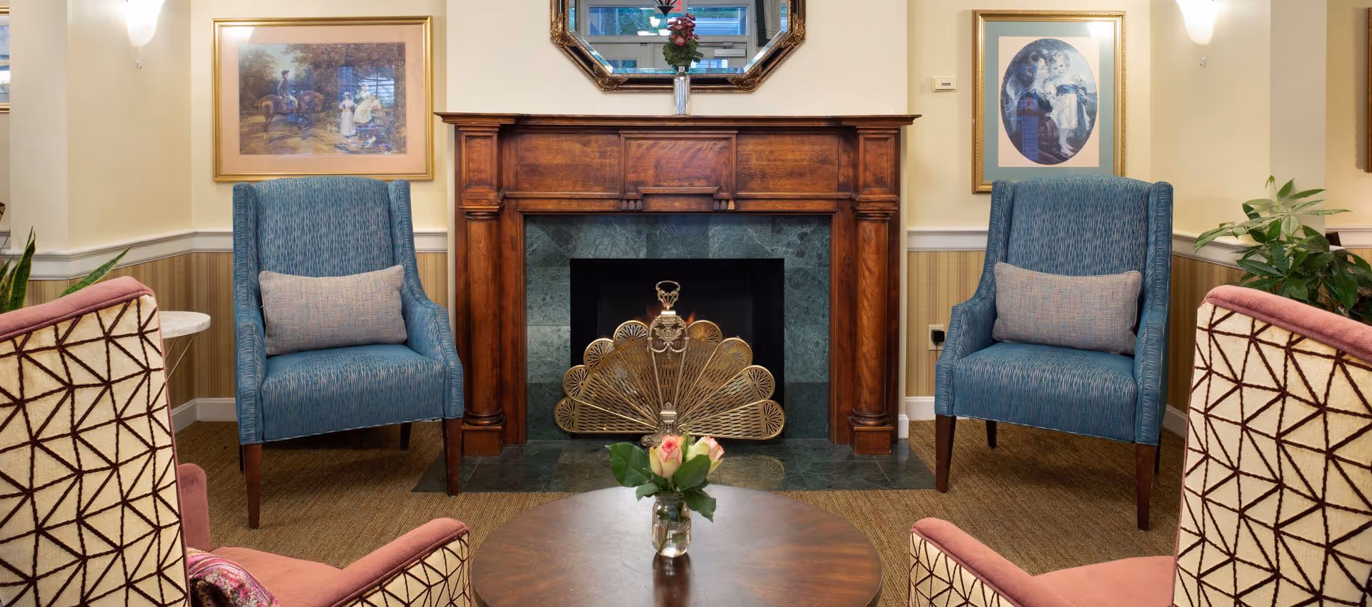 Cozy lounge with patterned and blue upholstered armchairs arranged around a round wooden table facing a wood-and-marble fireplace.