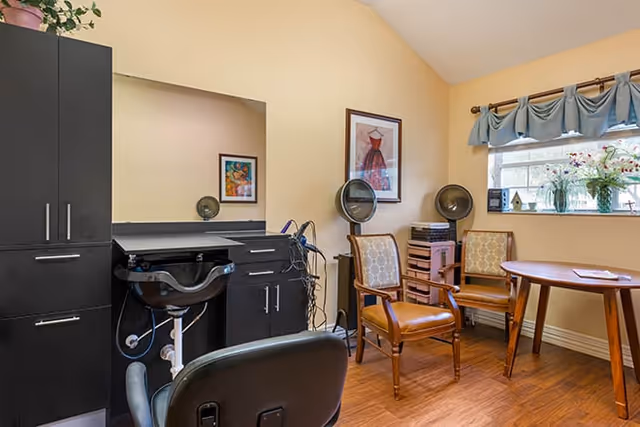 A cozy salon room with a styling station, shampoo sink, salon chairs, hair dryers, cabinets, and a small table by a window.