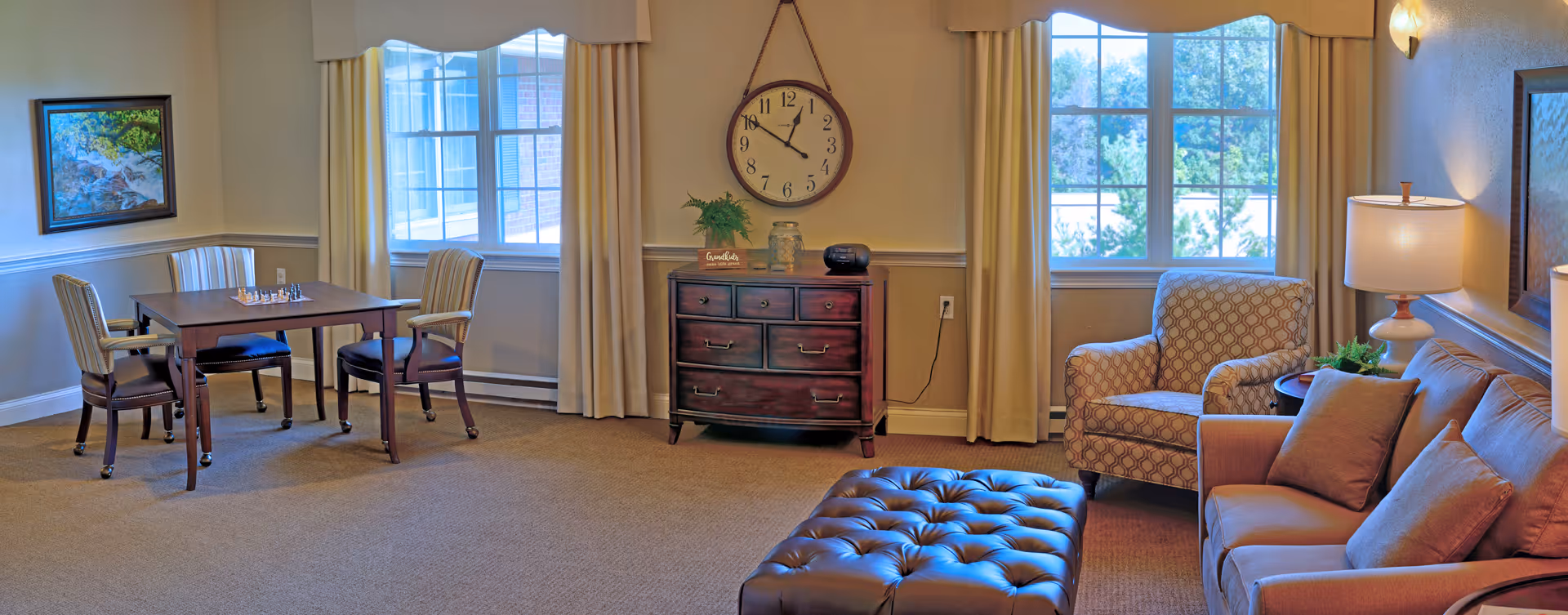 A cozy living room area with a brown leather sofa, a patterned armchair, a large tufted ottoman, and a wooden side table with a lamp. There is a wooden dresser with a round wall clock above it, and a small table with four chairs near a window. The room has beige walls, carpeted floor, and large windows with cream-colored curtains letting in natural light.