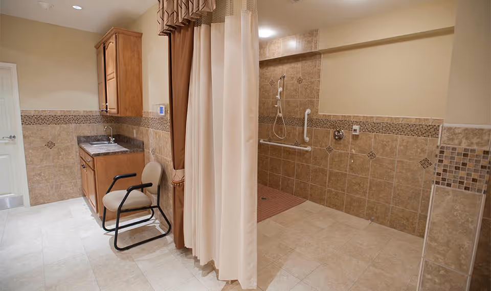 Accessible tiled bathroom with a roll-in shower, grab bars, sink cabinet and a chair separated by a privacy curtain.